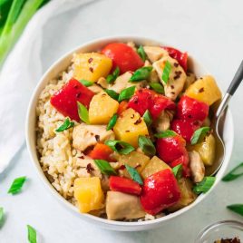 A bowl of brown rice topped with chicken, red bell peppers, pineapple, and chopped green onions, with a spoon.