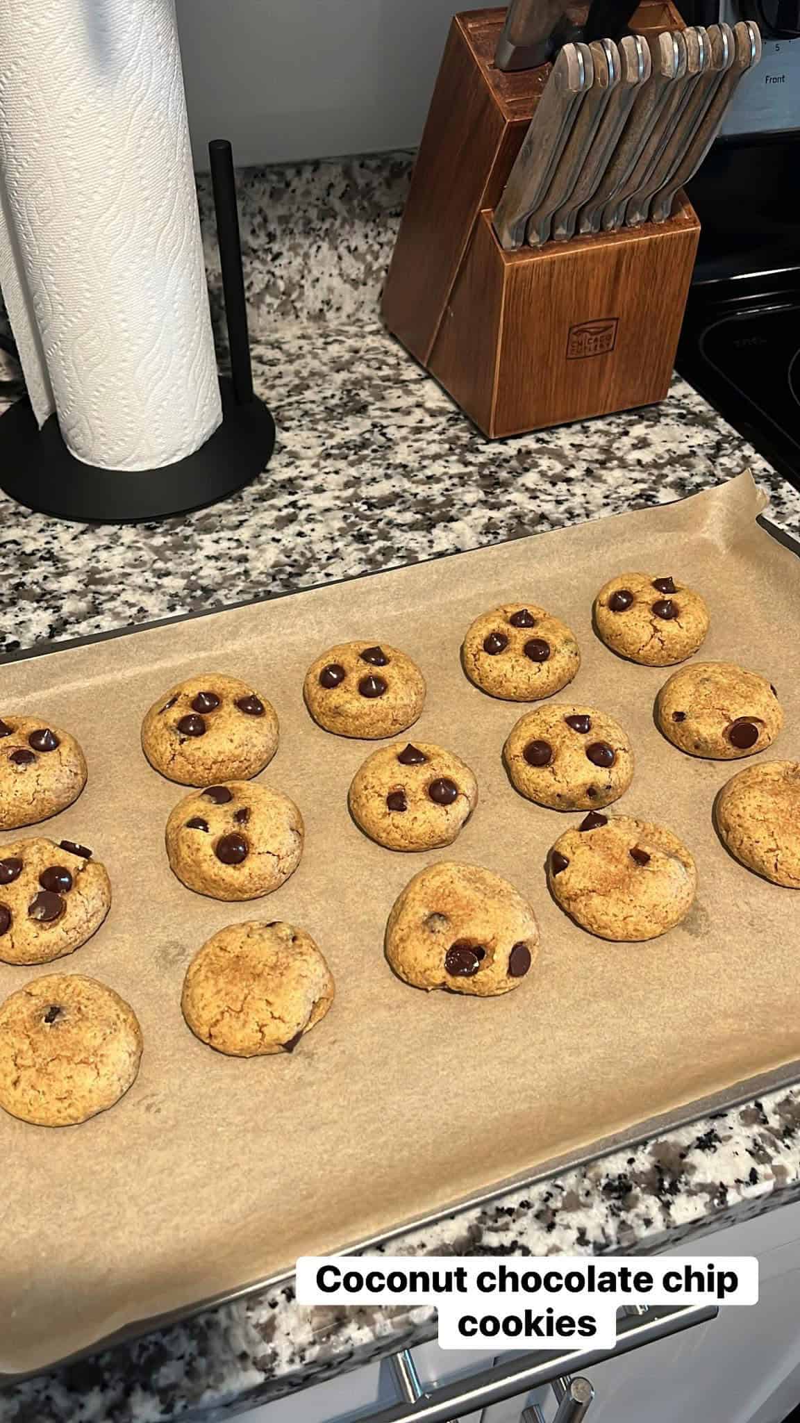 A baking tray with coconut chocolate chip cookies on parchment paper, on a kitchen counter near a knife block and paper towels.