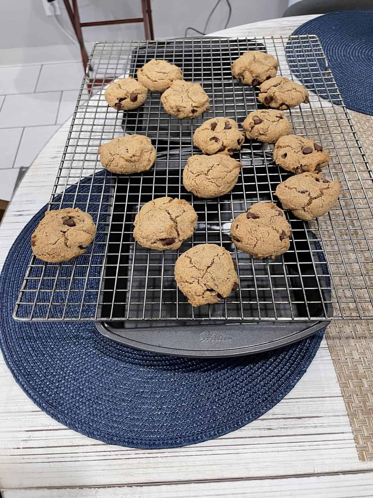 A cooling rack with chocolate chip cookies resting on it is placed over a gray round baking tray. The setup is on a white table with a blue placemat beneath.