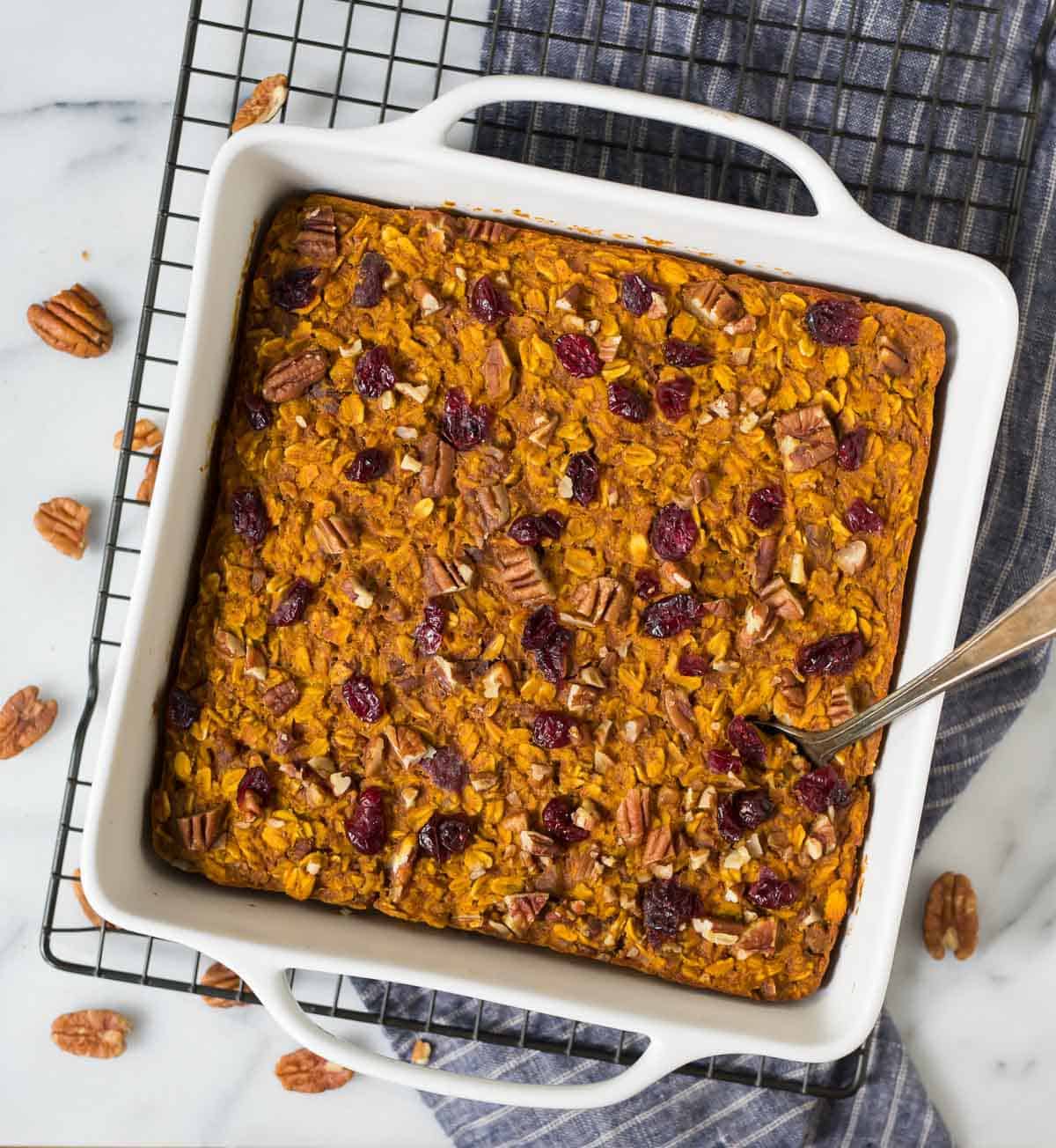 A pumpkin baked oatmeal with cranberries and pecans in a white dish on a cooling rack, with a spoon inside.