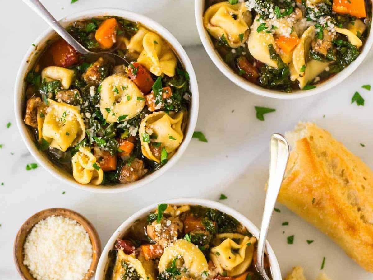 Three bowls of tortellini soup with vegetables and herbs, served with grated cheese and pieces of bread on a marble surface.
