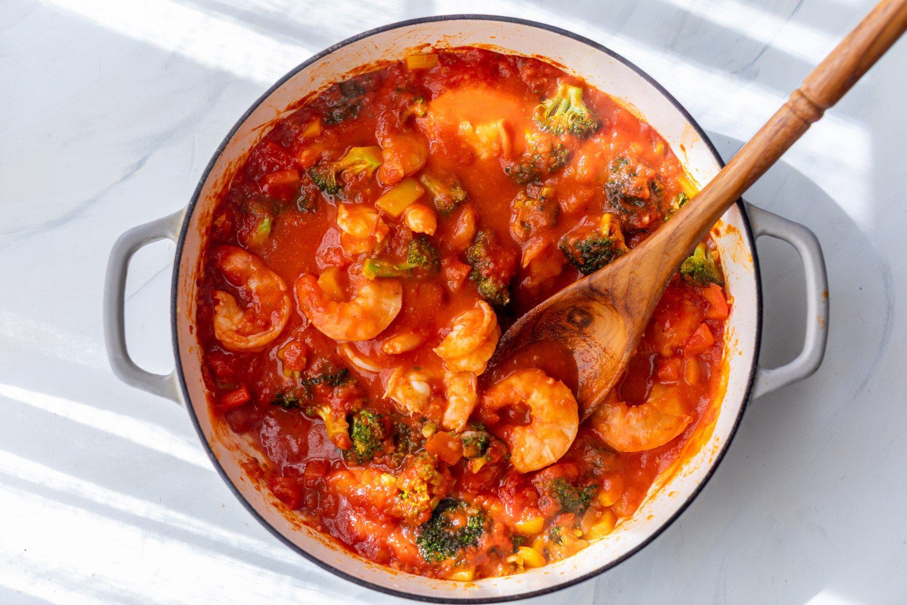 A pot of shrimp and vegetables in tomato sauce with a wooden spoon resting inside.