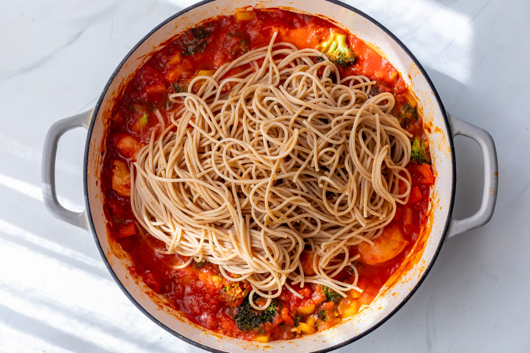 Spaghetti on top of vegetable tomato sauce in a white pot, ready to be mixed together for spicy shrimp pasta.