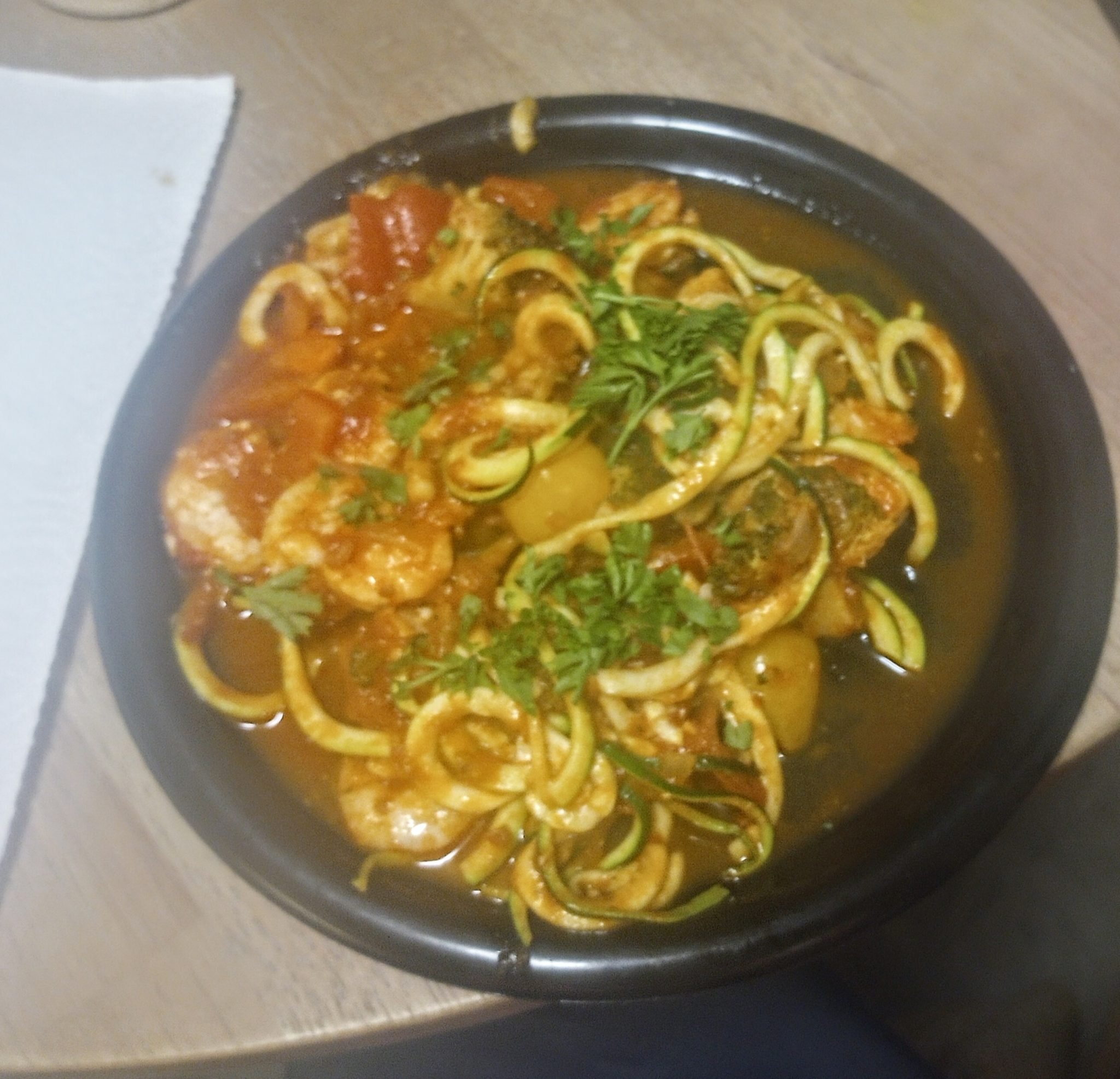 A bowl of shrimp and vegetable noodle soup topped with chopped herbs on a wooden table.