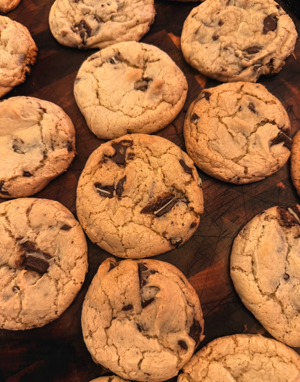 A batch of freshly baked chocolate chip cookies on a wooden surface. The cookies are golden brown with visible chunks of chocolate.