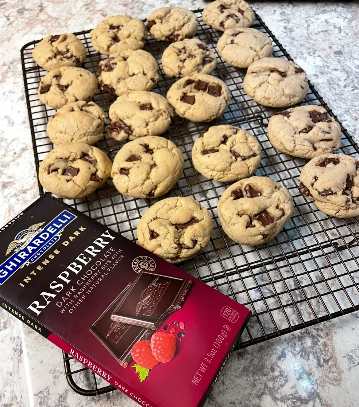 Freshly baked cookies with chunks of chocolate are cooling on a wire rack beside a Ghirardelli Raspberry Dark Chocolate bar. The countertop beneath is light-colored with a subtle pattern.