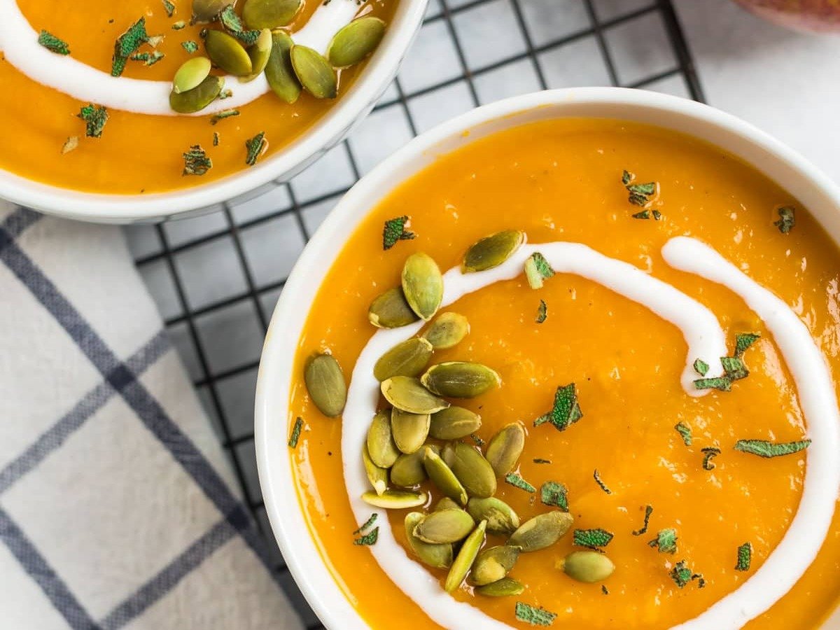 Two bowls of orange soup topped with cream, pumpkin seeds, and herbs, next to an apple and a checkered napkin.