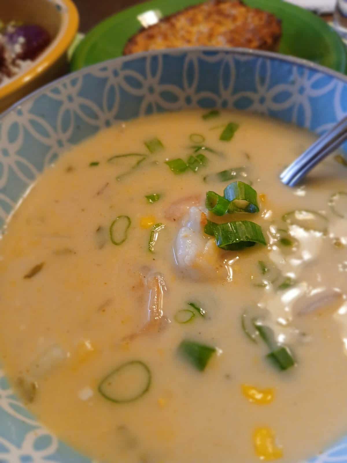A bowl of creamy soup with corn, green onions, and a spoon, with bread in the background.