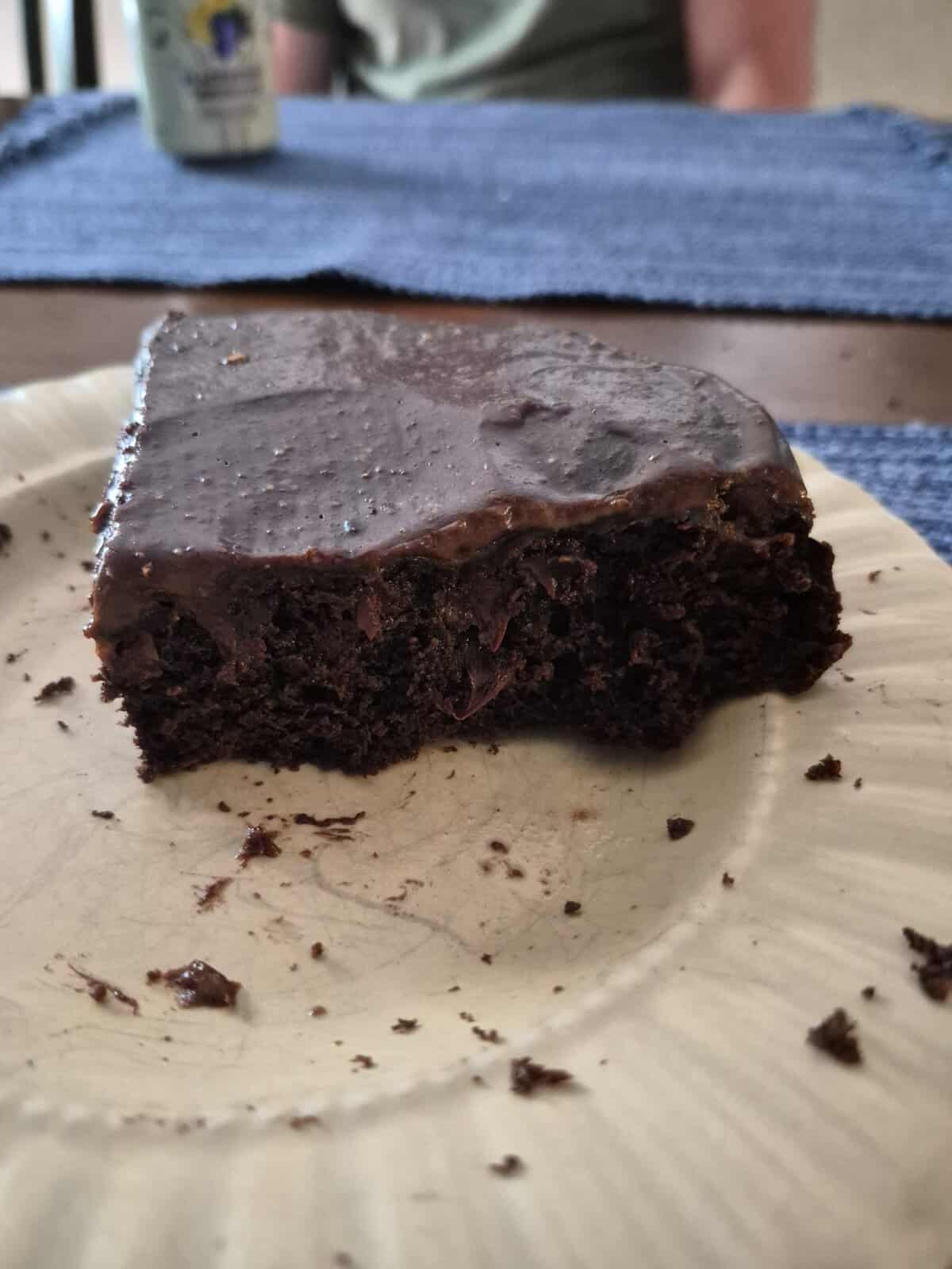 A partially eaten slice of chocolate cake with frosting sits on a beige plate; blue placemats are in the background.