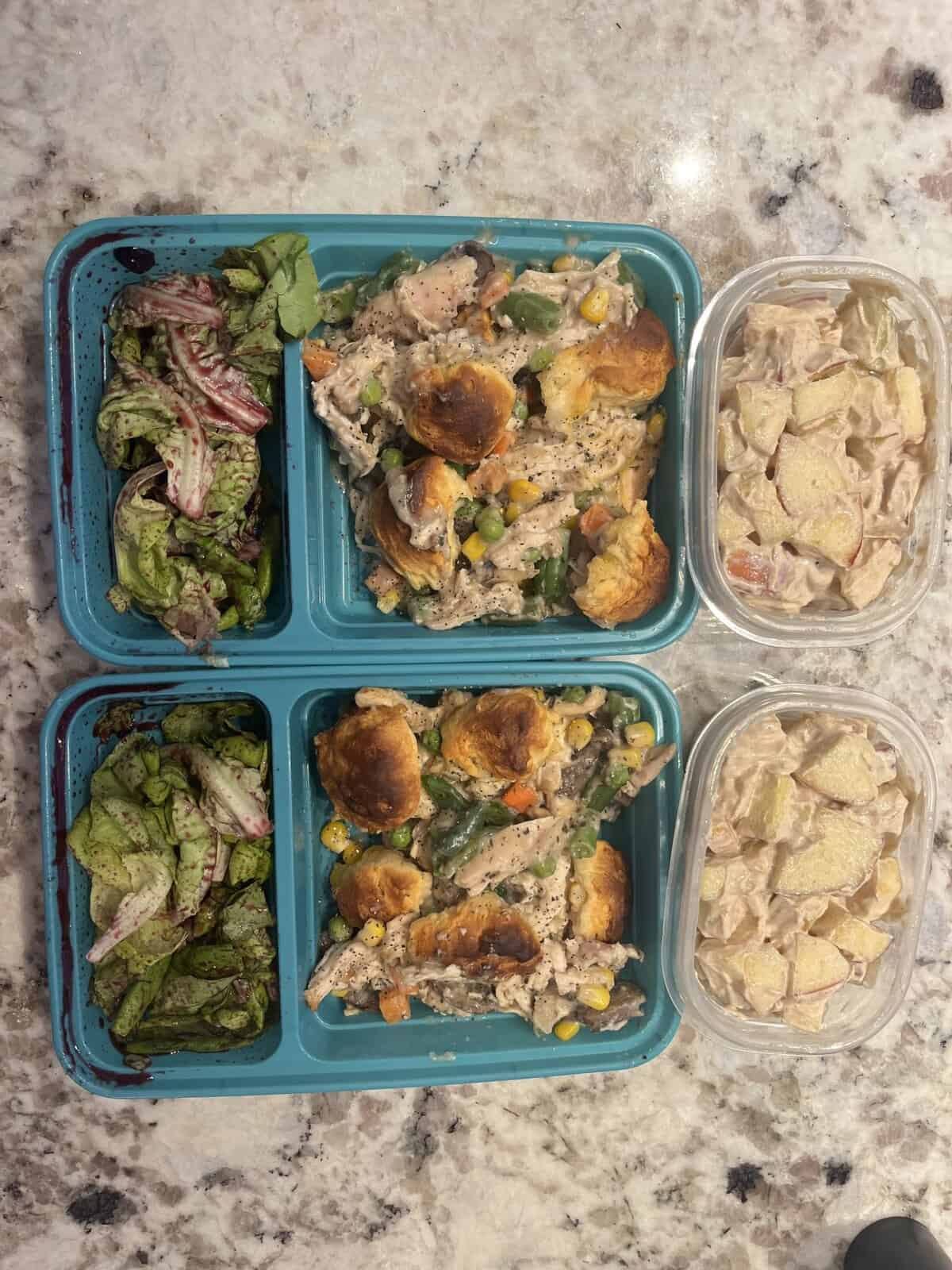 Two blue meal prep trays with salad and casserole, plus two containers of creamy pasta with potatoes on a marble counter.