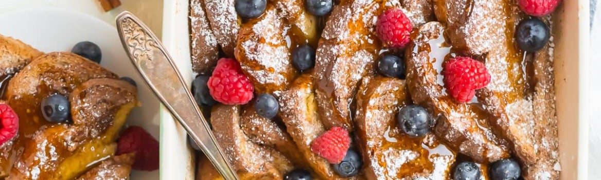 A baked French toast casserole topped with powdered sugar, fresh raspberries, and blueberries, with a serving spoon inside.