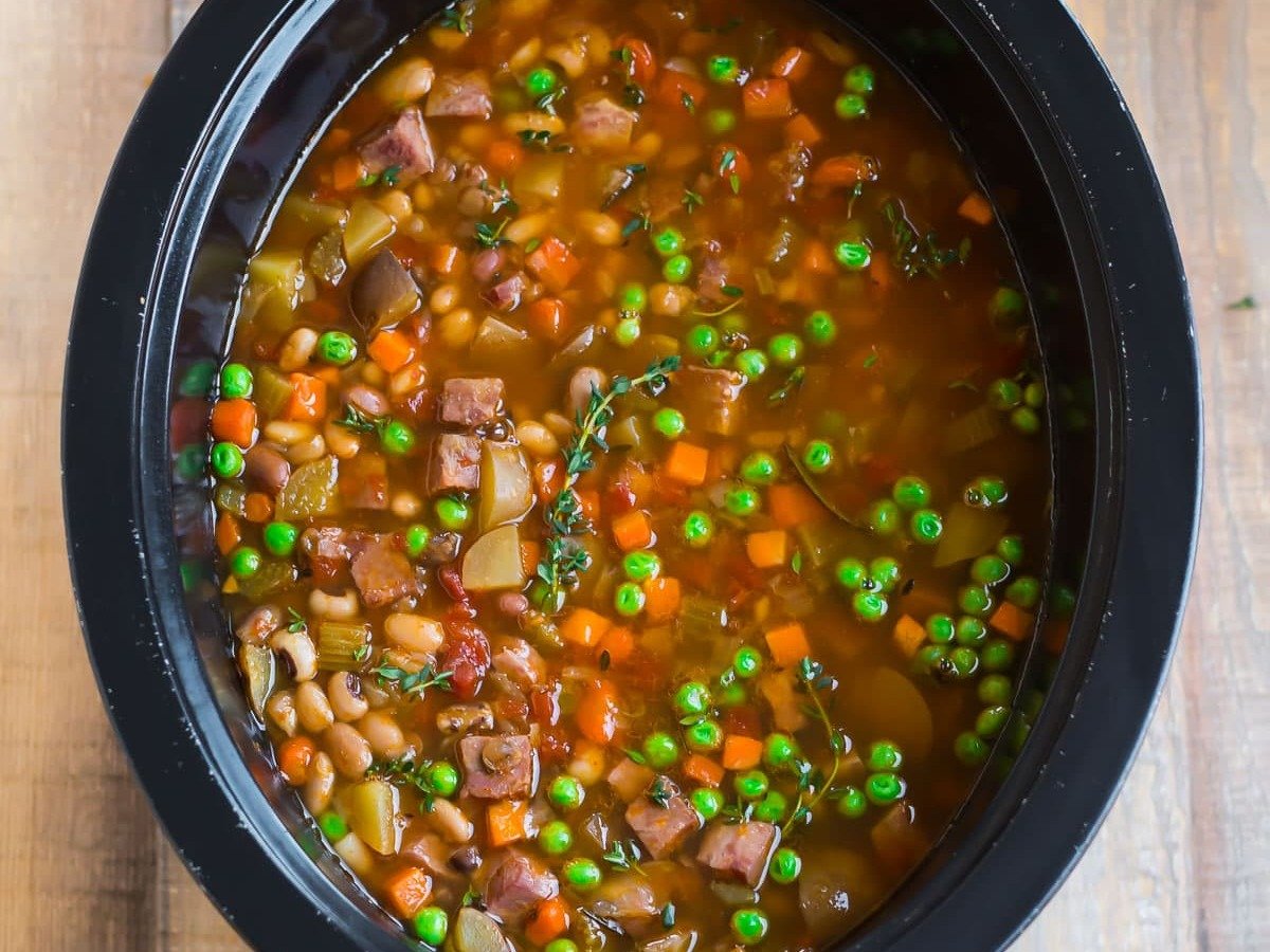 Slow cooker filled with vegetable and bean soup, including peas, carrots, beans, and diced meat in broth.