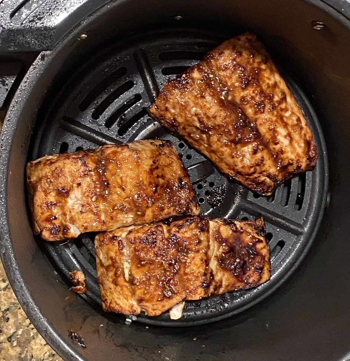 Three glazed salmon fillets cooking in an air fryer basket with a dark nonstick coating.