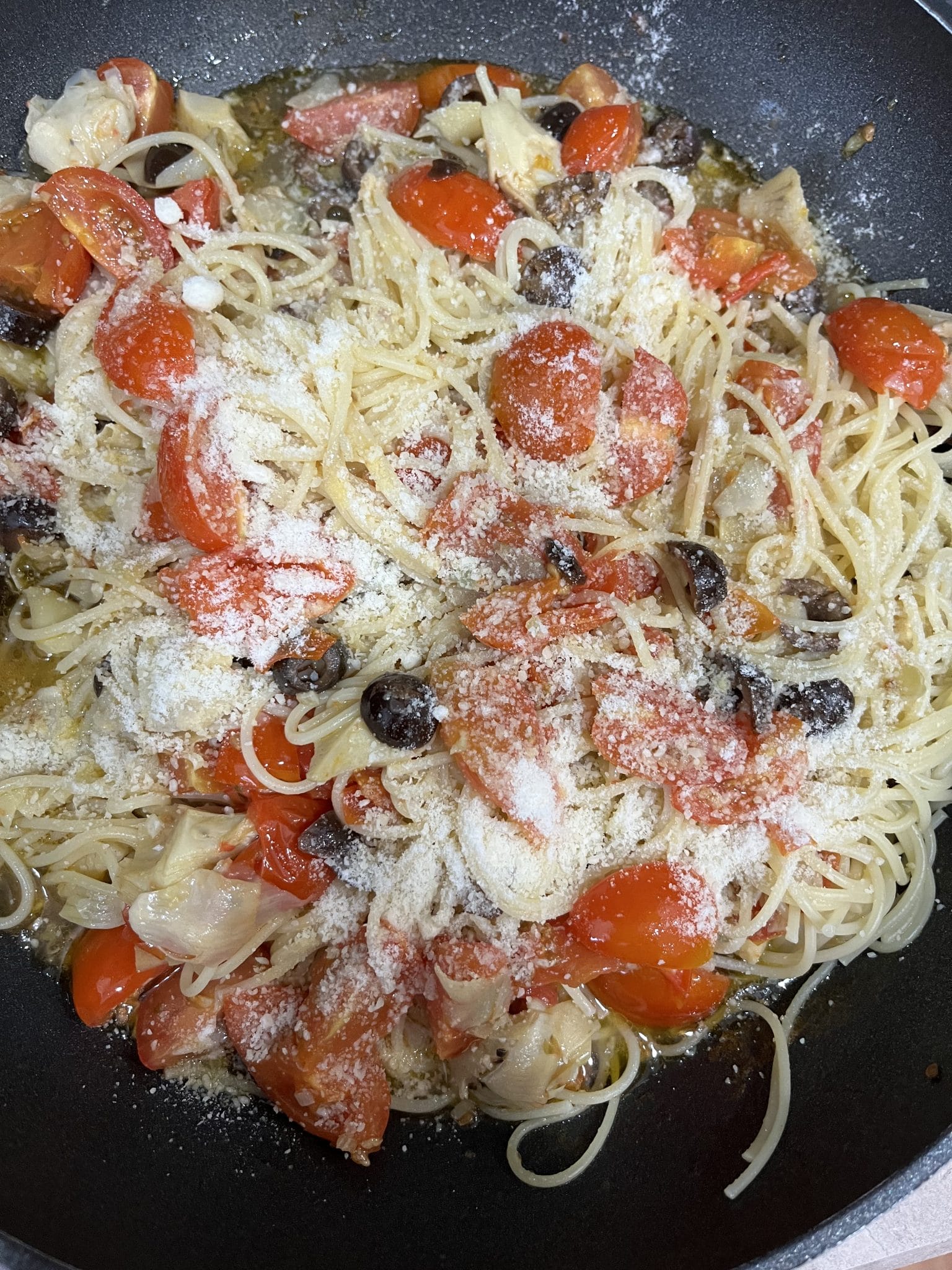 A pan of cooked spaghetti mixed with cherry tomatoes, black olives, garlic, and topped with grated parmesan cheese.
