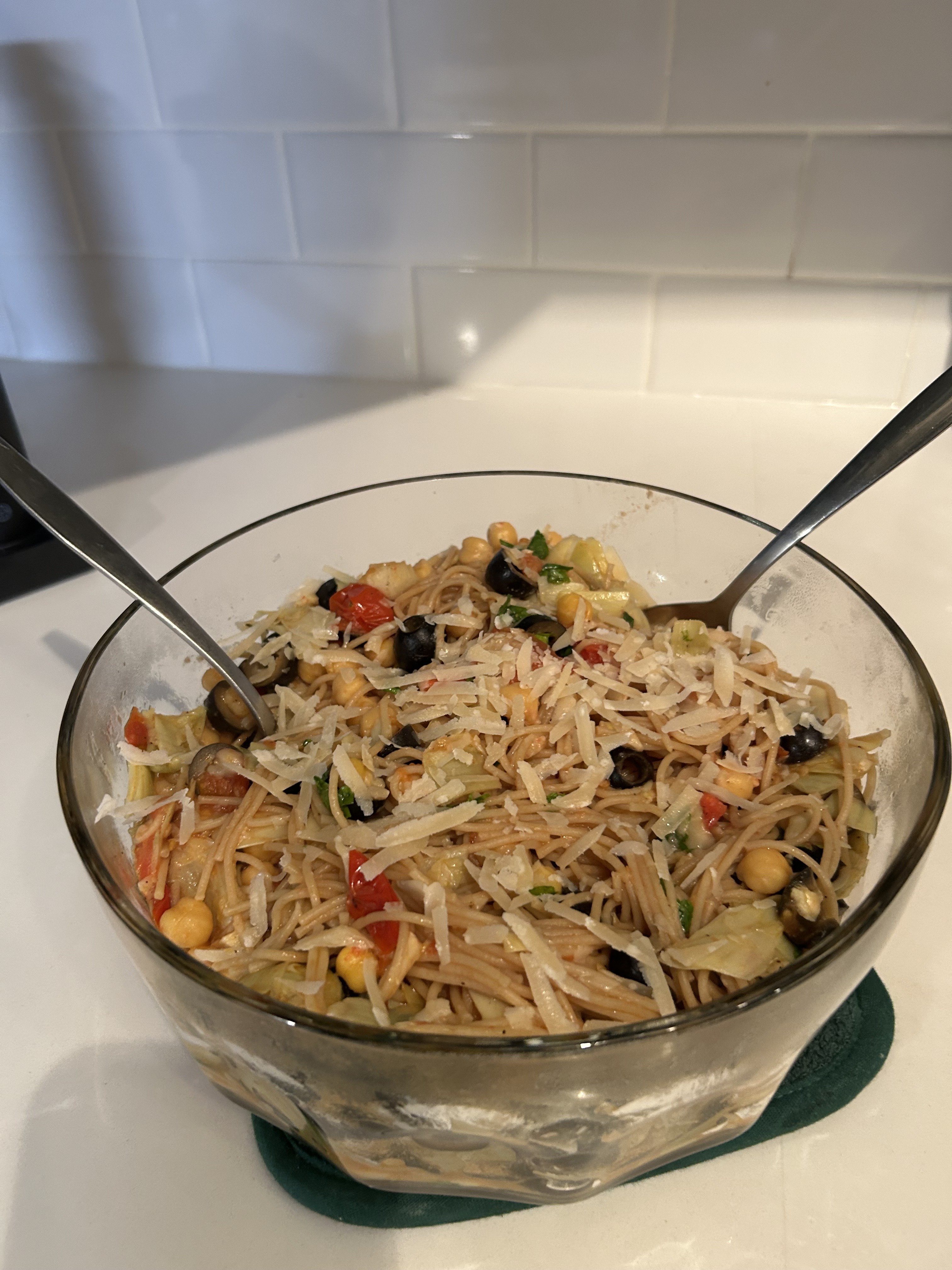 A glass bowl of spaghetti salad with vegetables, olives, chickpeas, and shredded cheese, with two serving utensils.