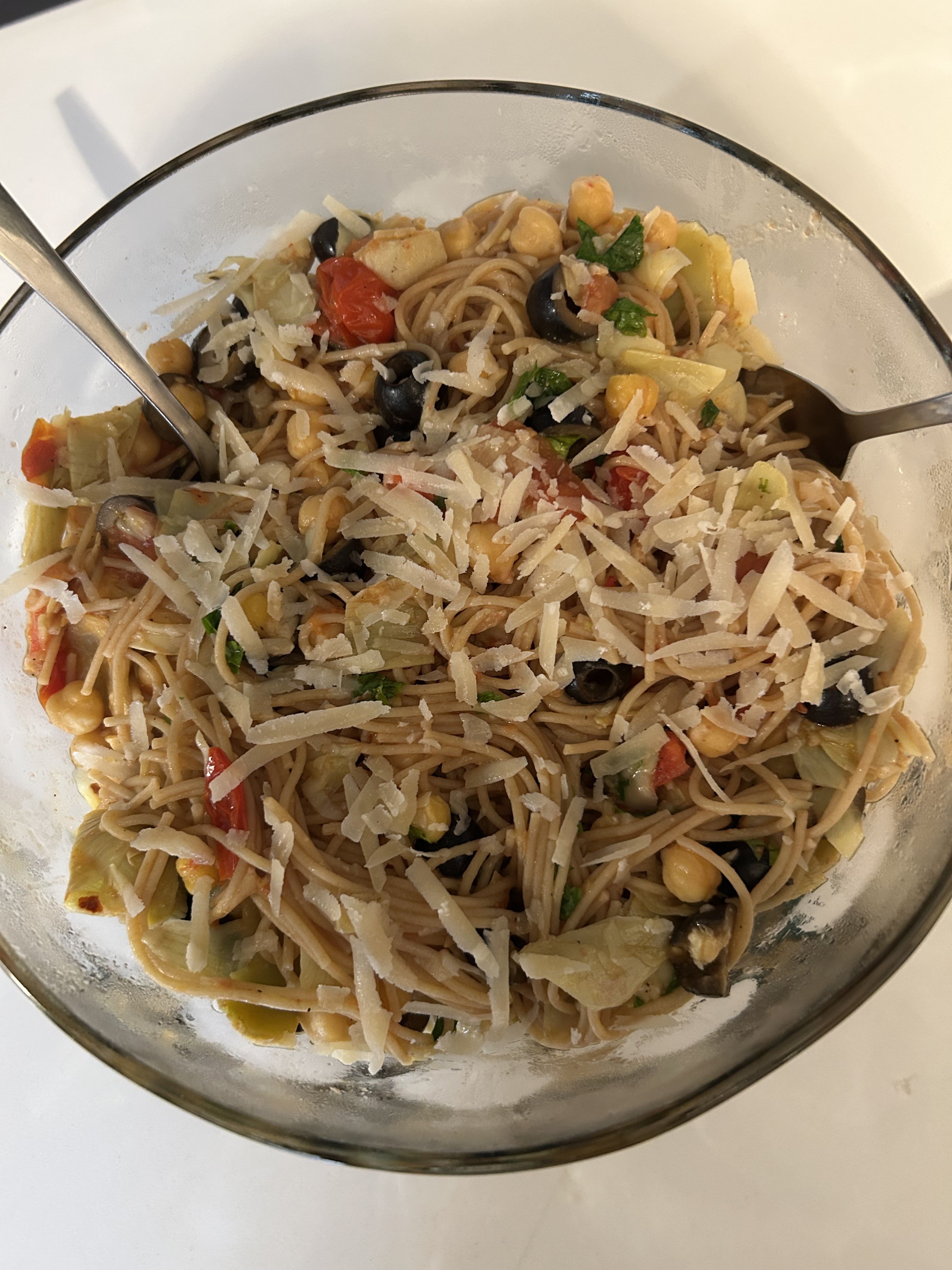 A bowl of pasta salad with vegetables, black olives, and shredded cheese, with two forks in the bowl.