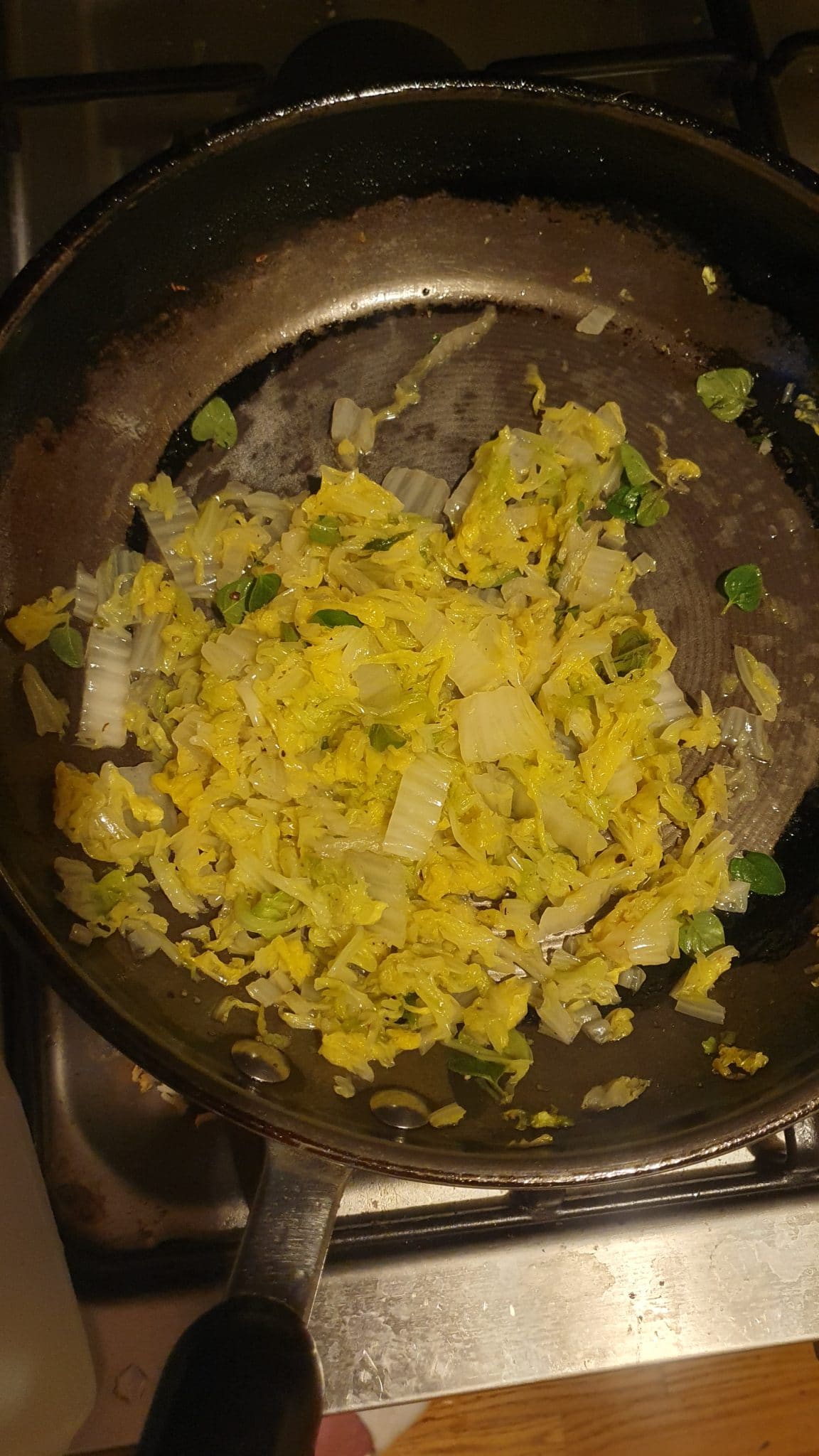Chopped cabbage, onions, and herbs sautéing in a black frying pan on a stove.