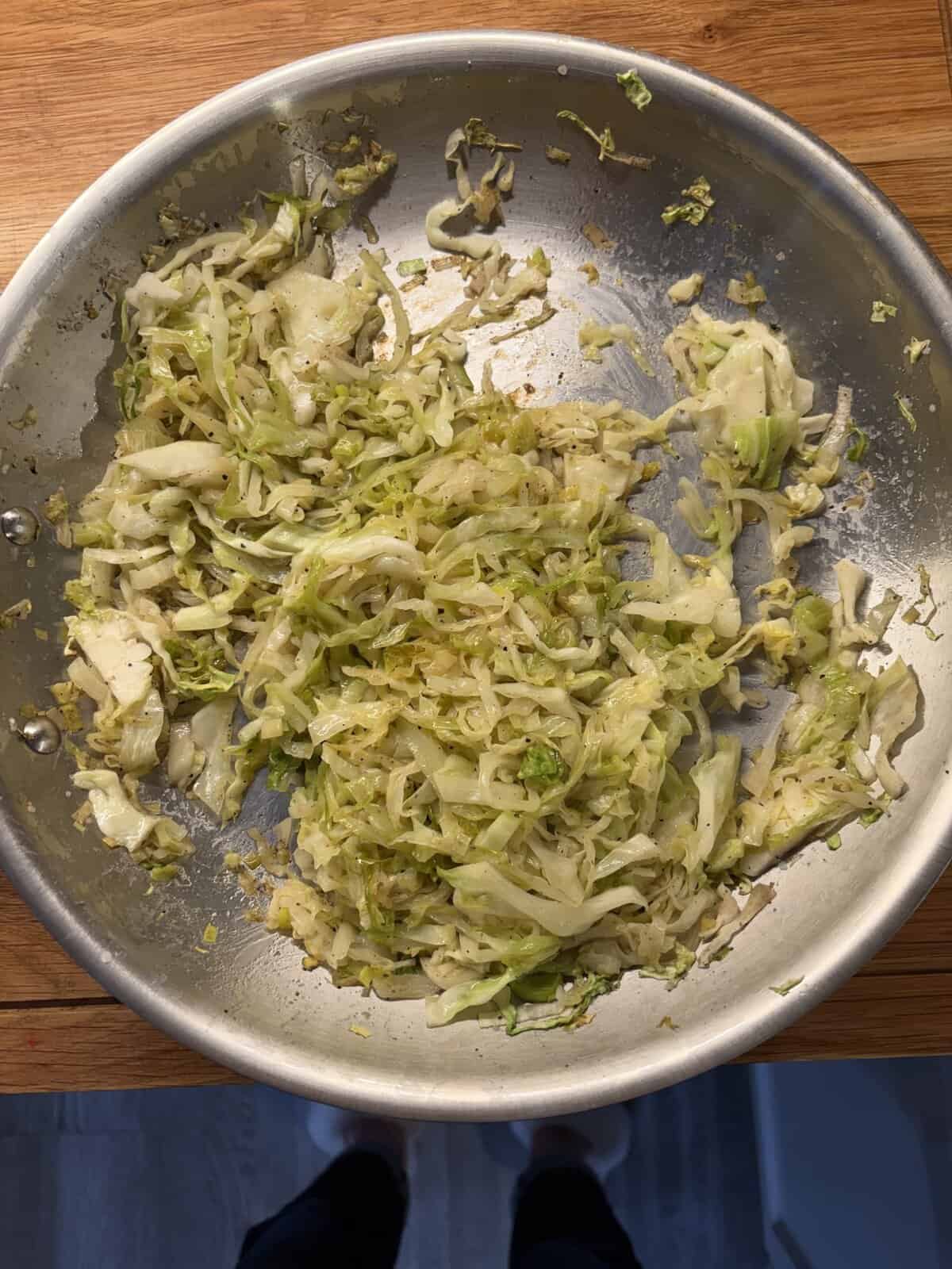 Shredded cabbage sautéing in a stainless steel pan on a wooden countertop.