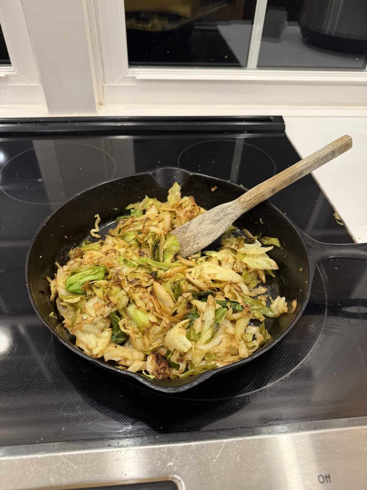 A cast iron skillet with sautéed cabbage and a wooden spatula rests on a stovetop in front of a window.