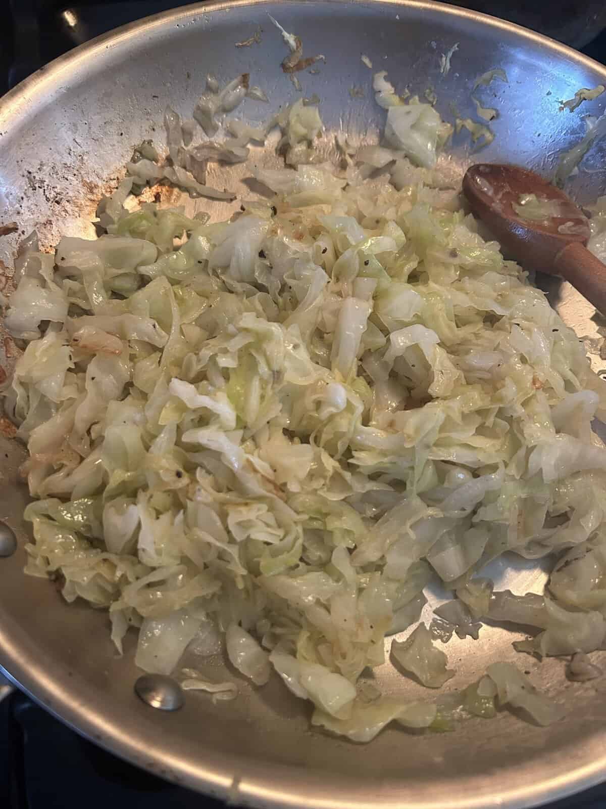 Shredded cabbage being sautéed in a stainless steel pan with a wooden spoon.