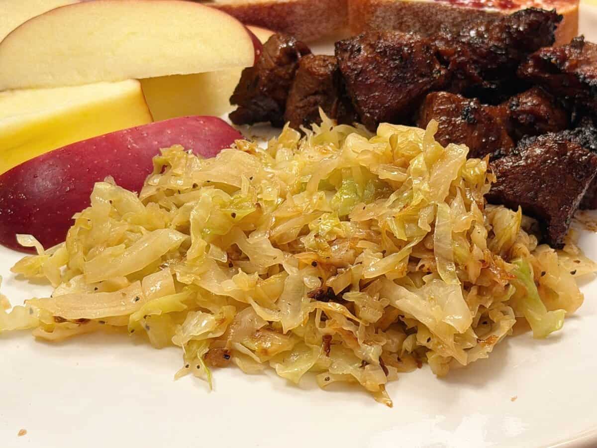 Shredded sautéed cabbage, apple slices, and pieces of dark grilled meat on a white plate.