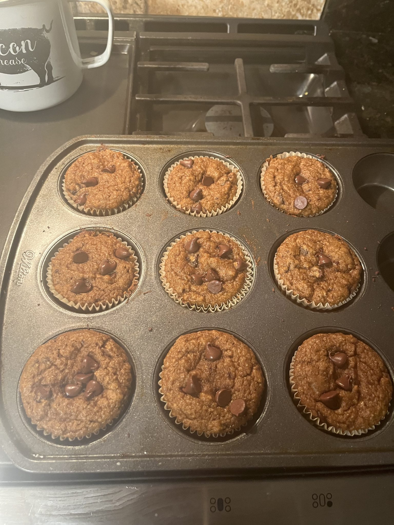 A muffin pan with nine baked chocolate chip muffins on a stovetop, with a white mug in the background.