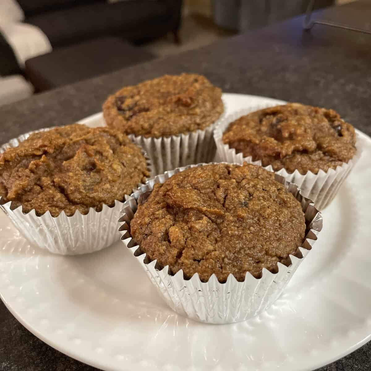 Four bran muffins in silver foil wrappers on a white plate, placed on a dark countertop.