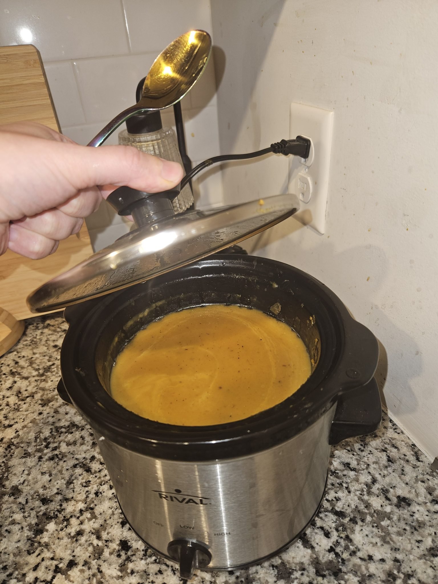 A hand lifts the lid of a slow cooker filled with creamy yellow soup on a kitchen counter.