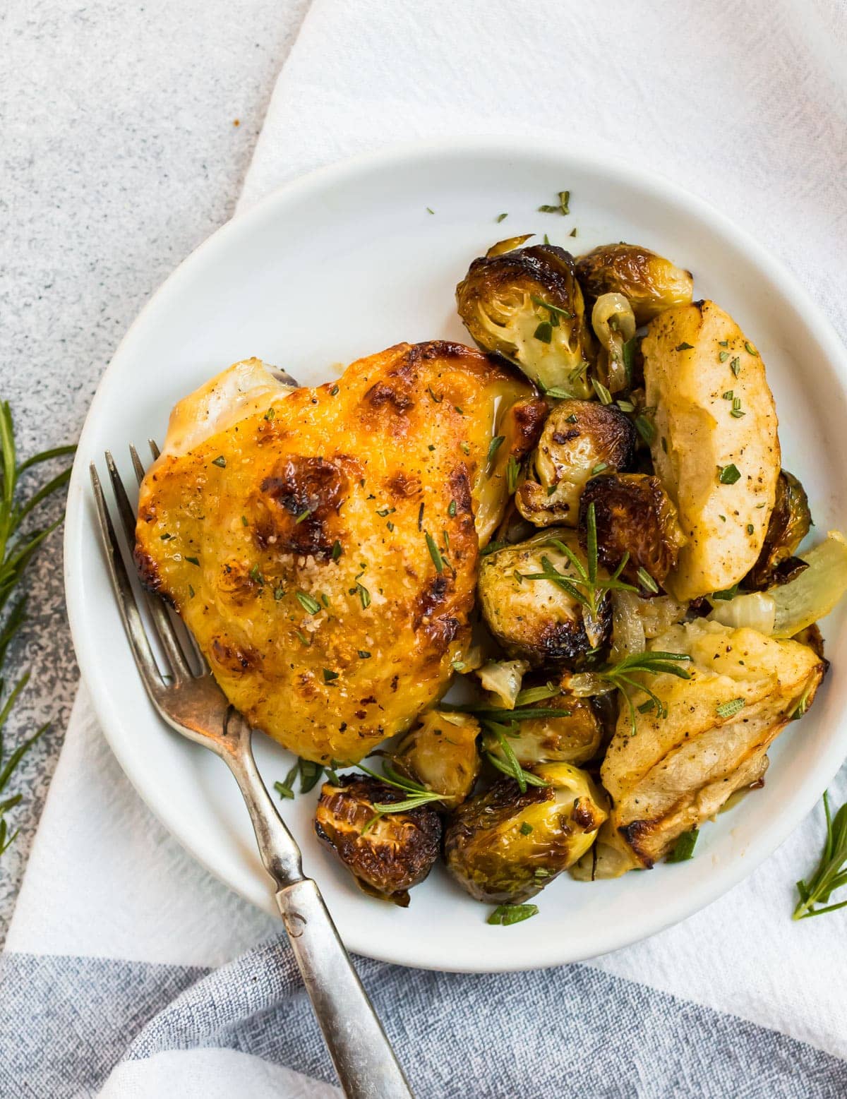 The BEST rosemary chicken thighs with honey mustard apples and Brussels sprouts on a white plate with fresh rosemary on top.