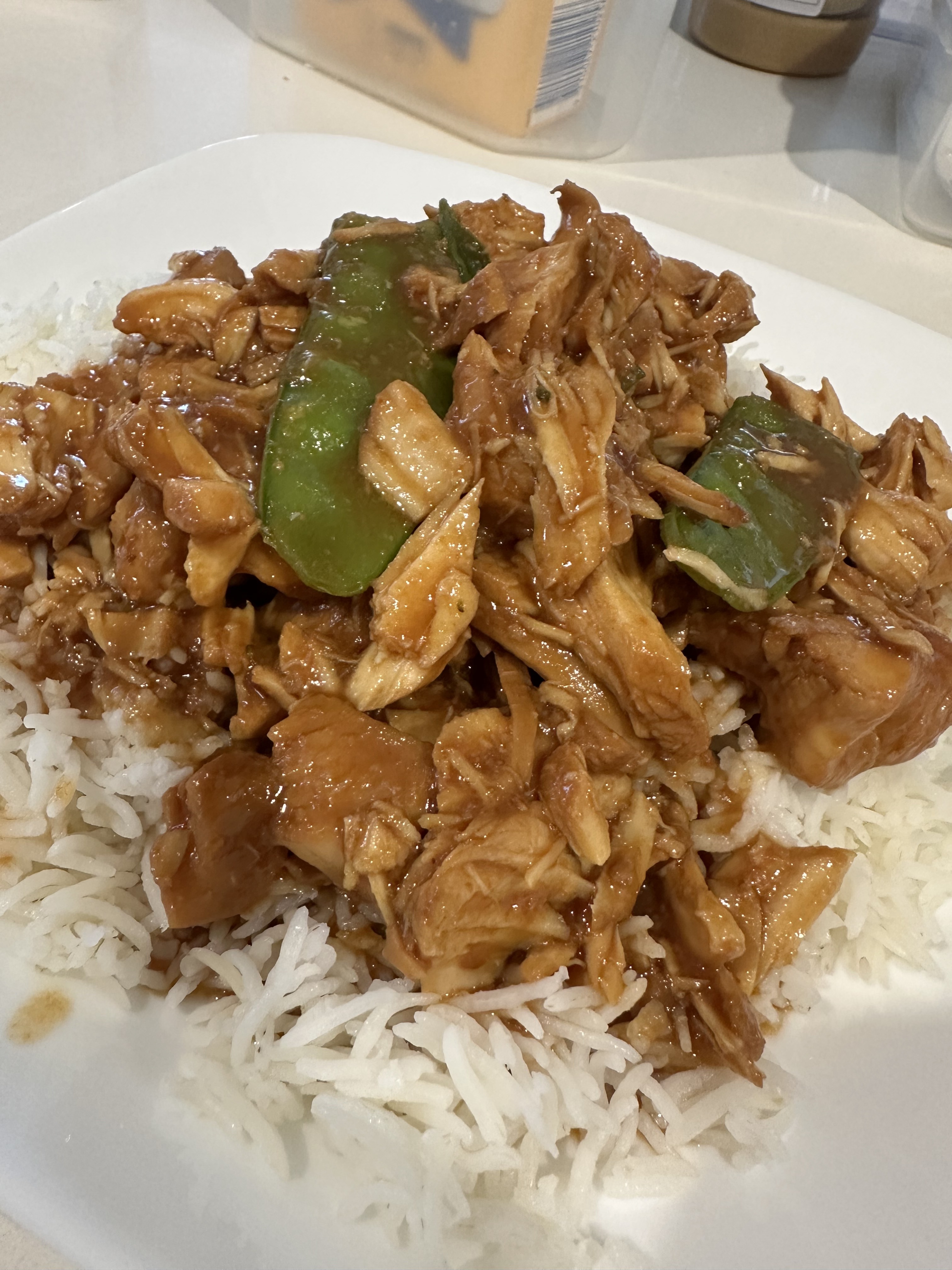 A plate of white rice topped with shredded chicken, green bell peppers, and a brown sauce, served on a white square dish.