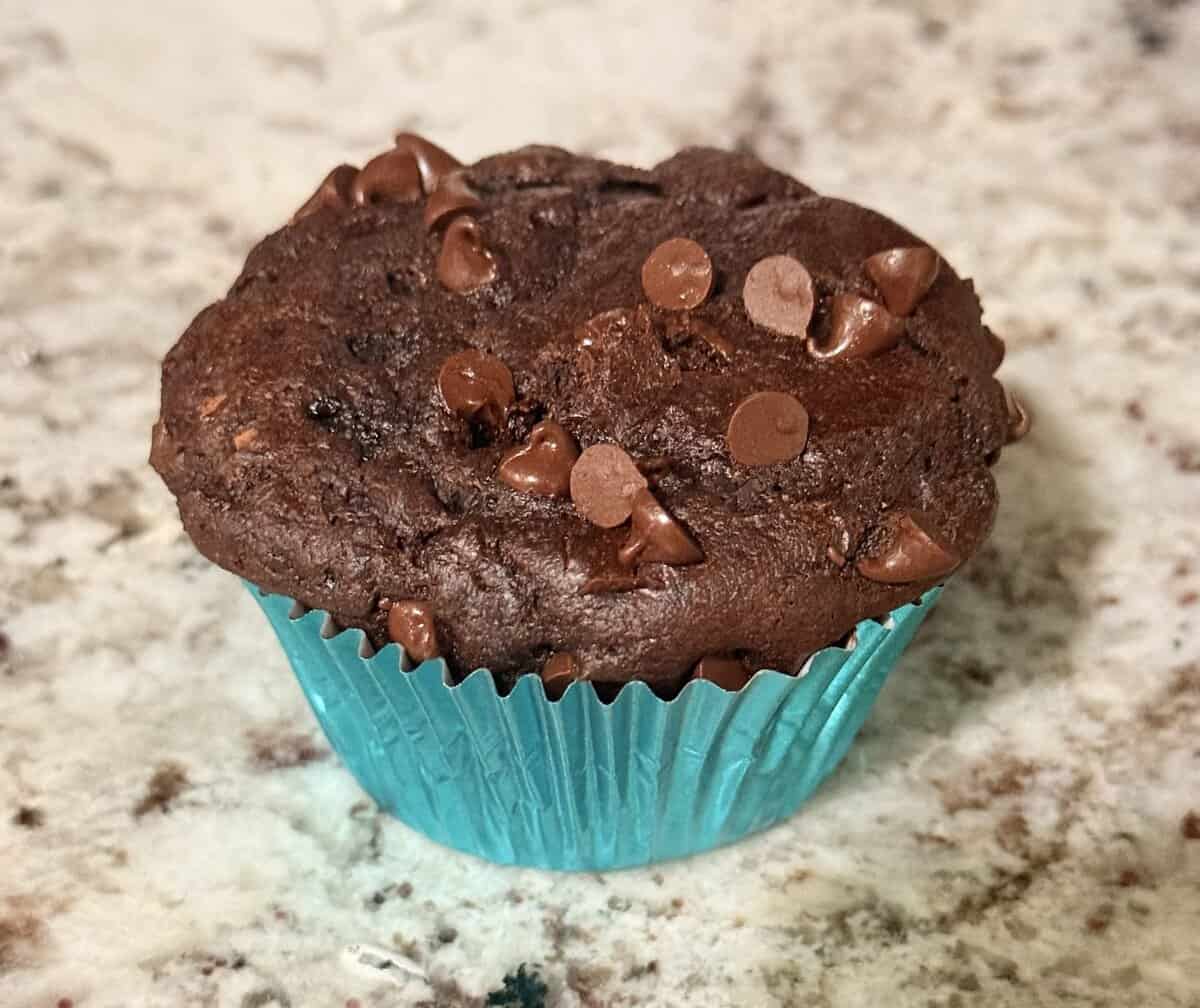 A chocolate muffin with chocolate chips in a blue foil wrapper sits on a speckled countertop.