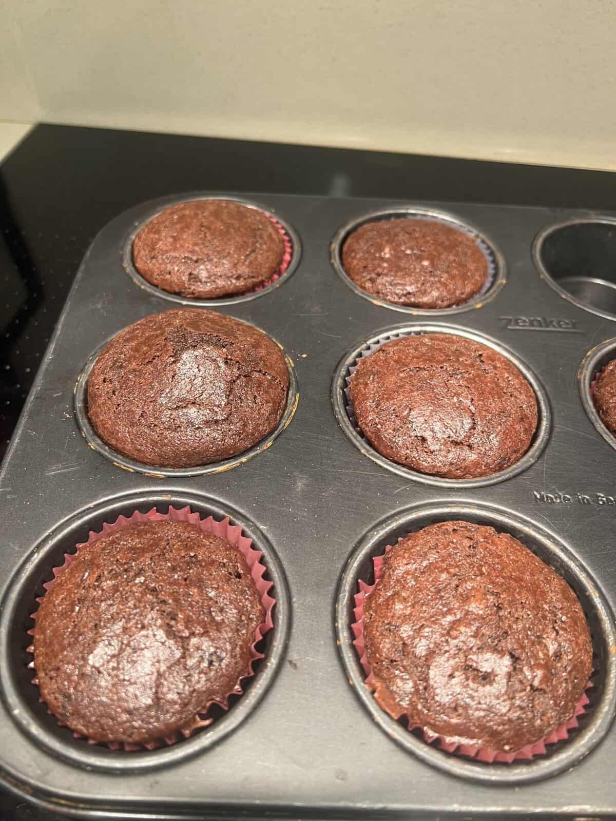 Freshly baked chocolate muffins in a six-cup muffin tin, with one muffin missing from a slot.
