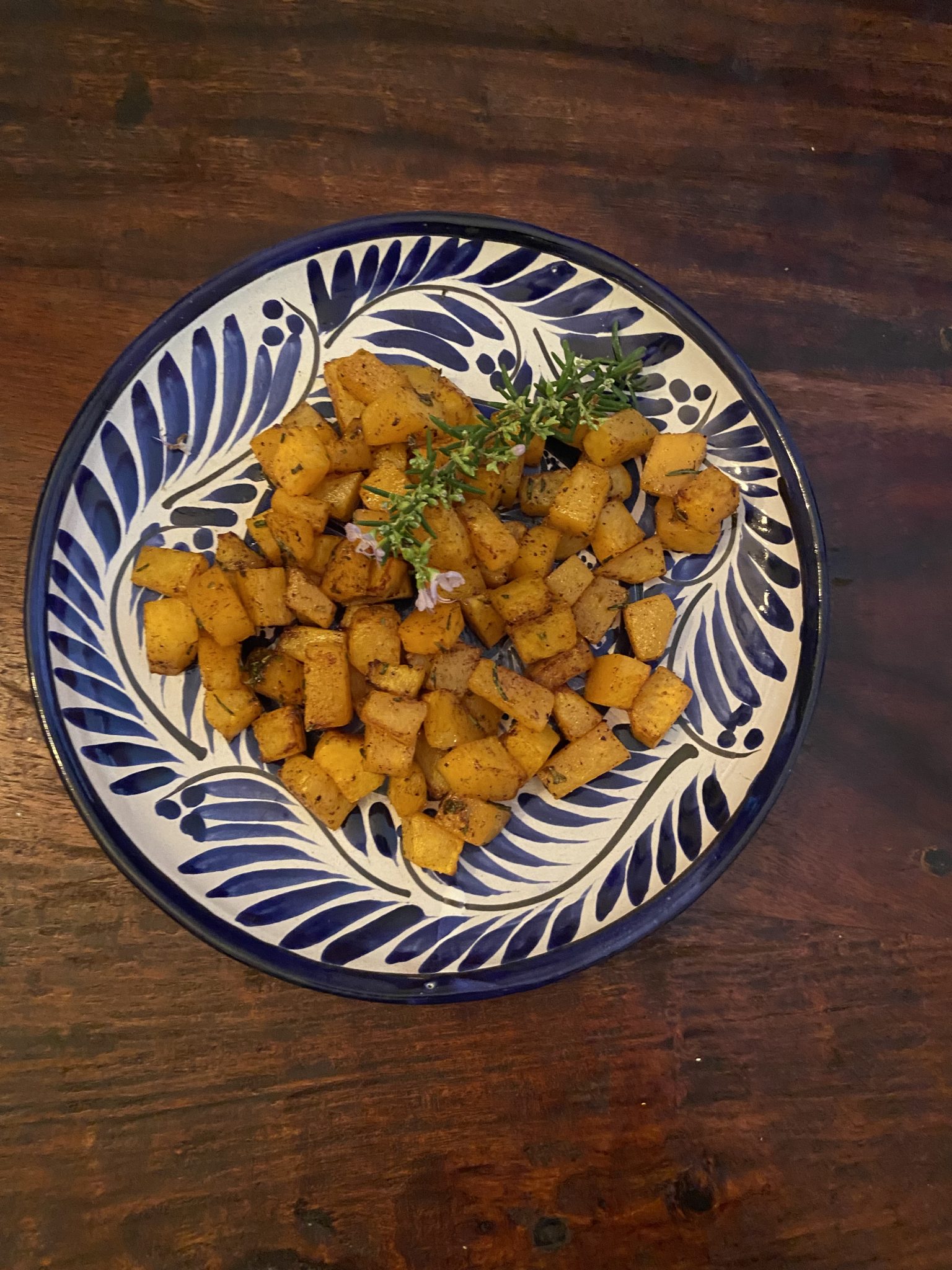 A decorative plate with cubed, roasted potatoes garnished with fresh herbs, on a dark wooden table.
