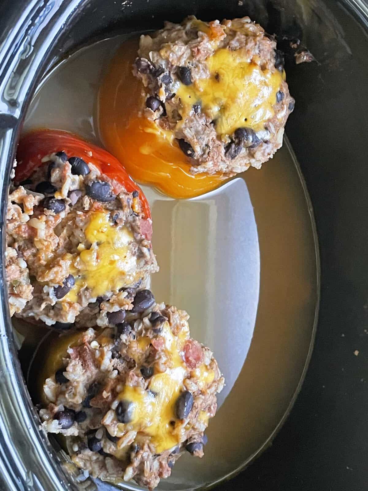 Three stuffed bell peppers with melted cheese and beans in a slow cooker with broth at the bottom.