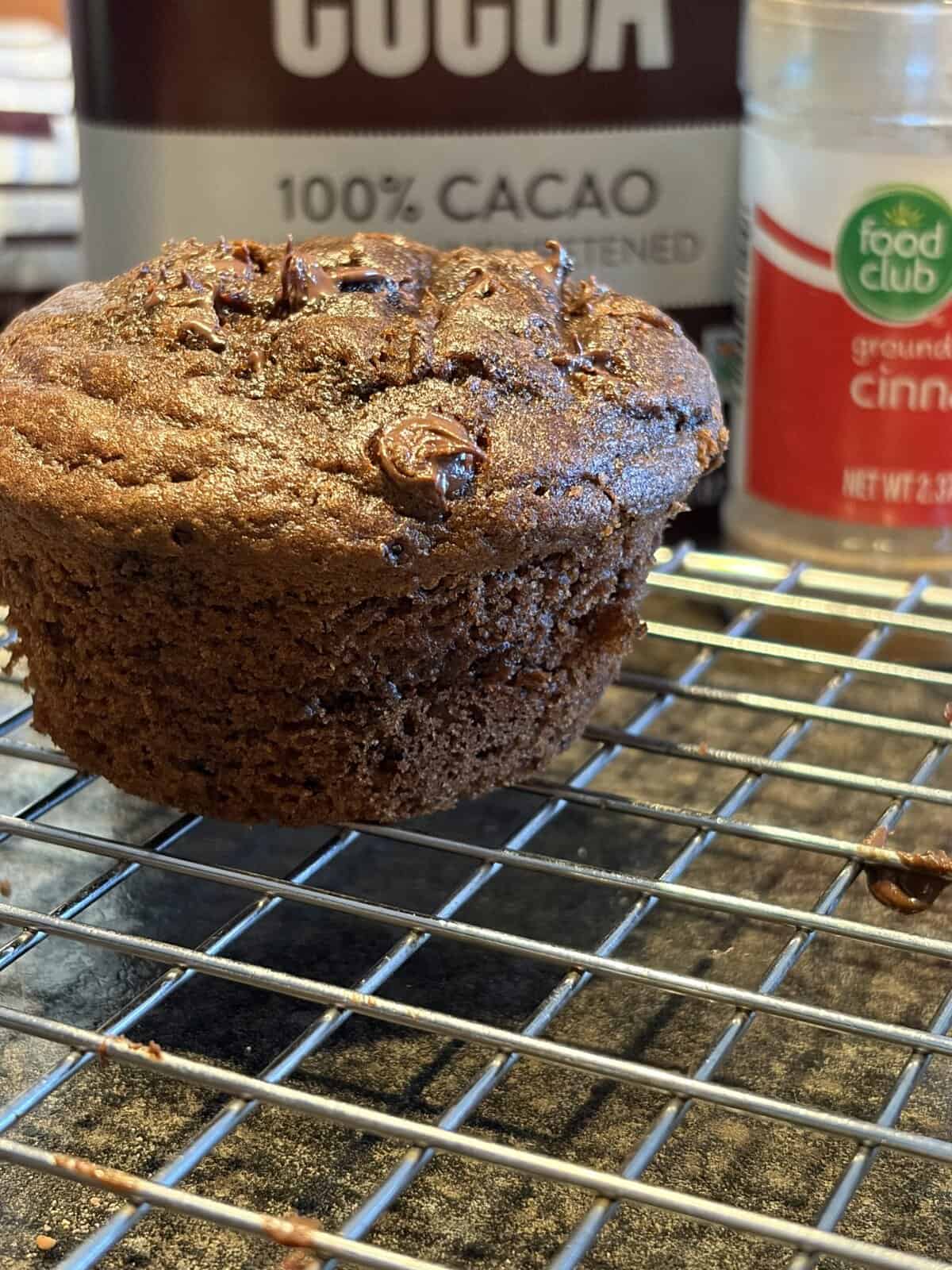 A chocolate muffin cools on a wire rack with cocoa and cinnamon containers in the background.