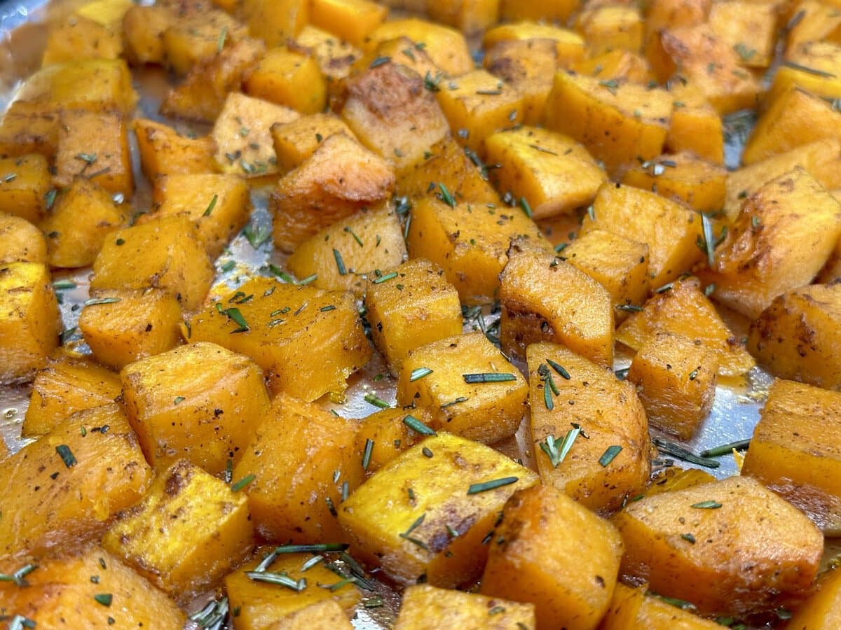 Close-up of roasted butternut squash cubes seasoned with herbs and spices on a baking sheet.