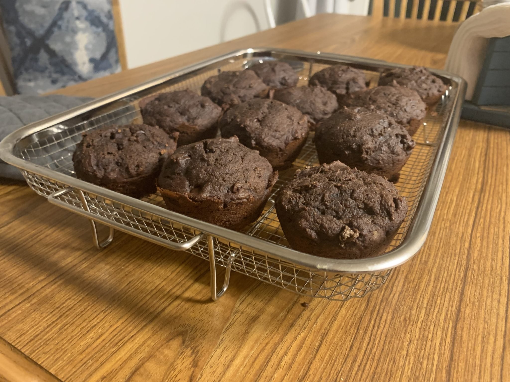 A wire rack holds twelve freshly baked chocolate muffins on a wooden table.