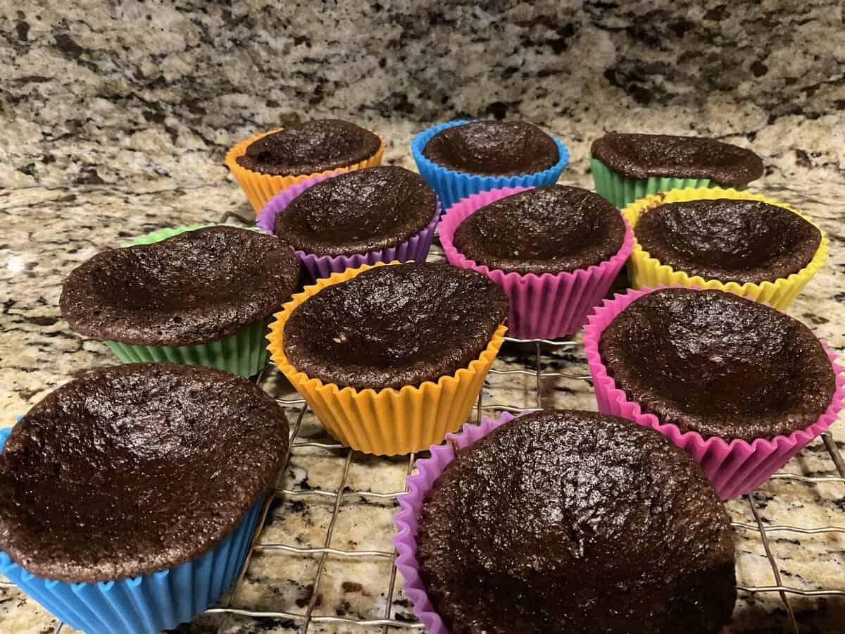 Twelve sunken chocolate cupcakes in colorful liners are cooling on a wire rack on a granite countertop.