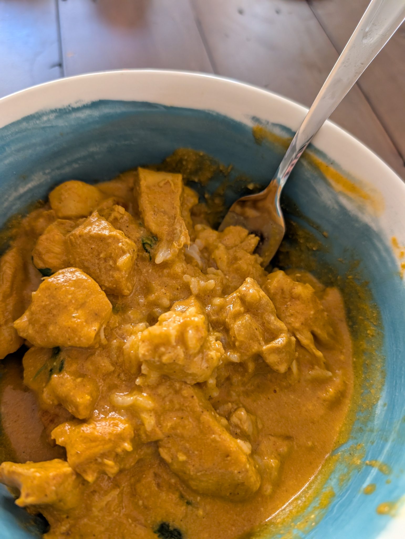 A bowl of creamy orange curry with chunks of chicken and rice, with a spoon resting inside the bowl.
