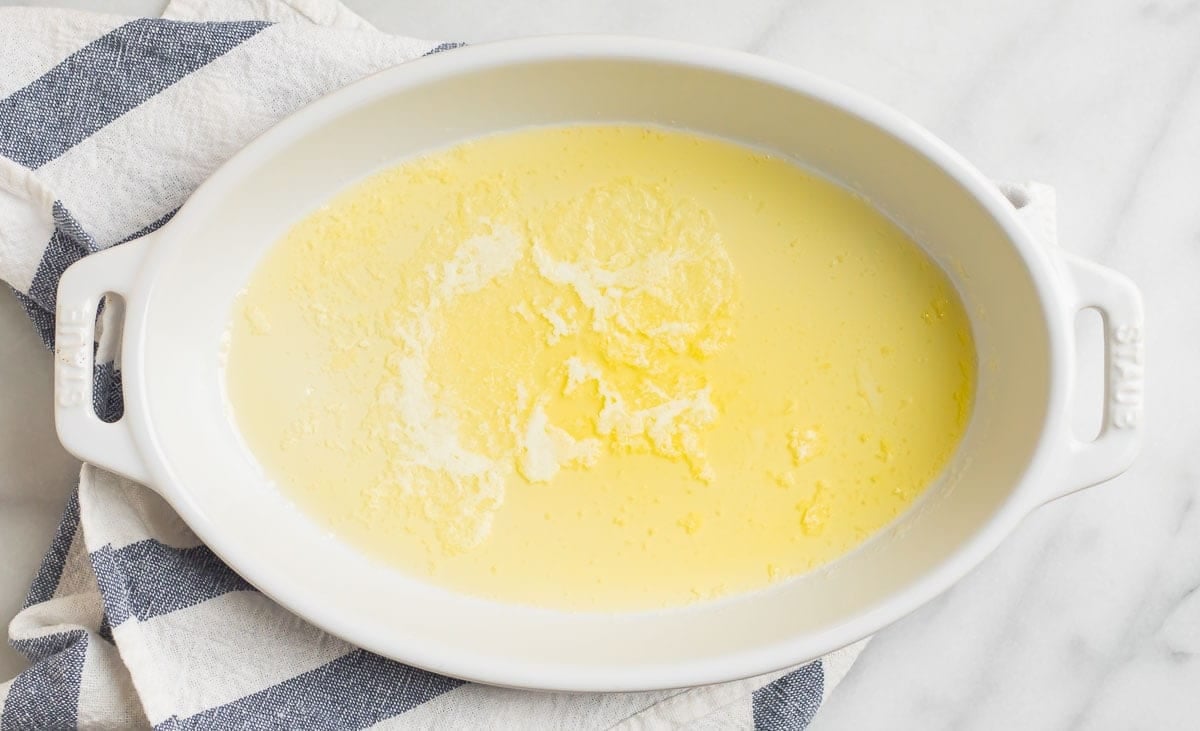A white oval baking dish containing melted butter, placed on a striped kitchen towel on a marble surface.
