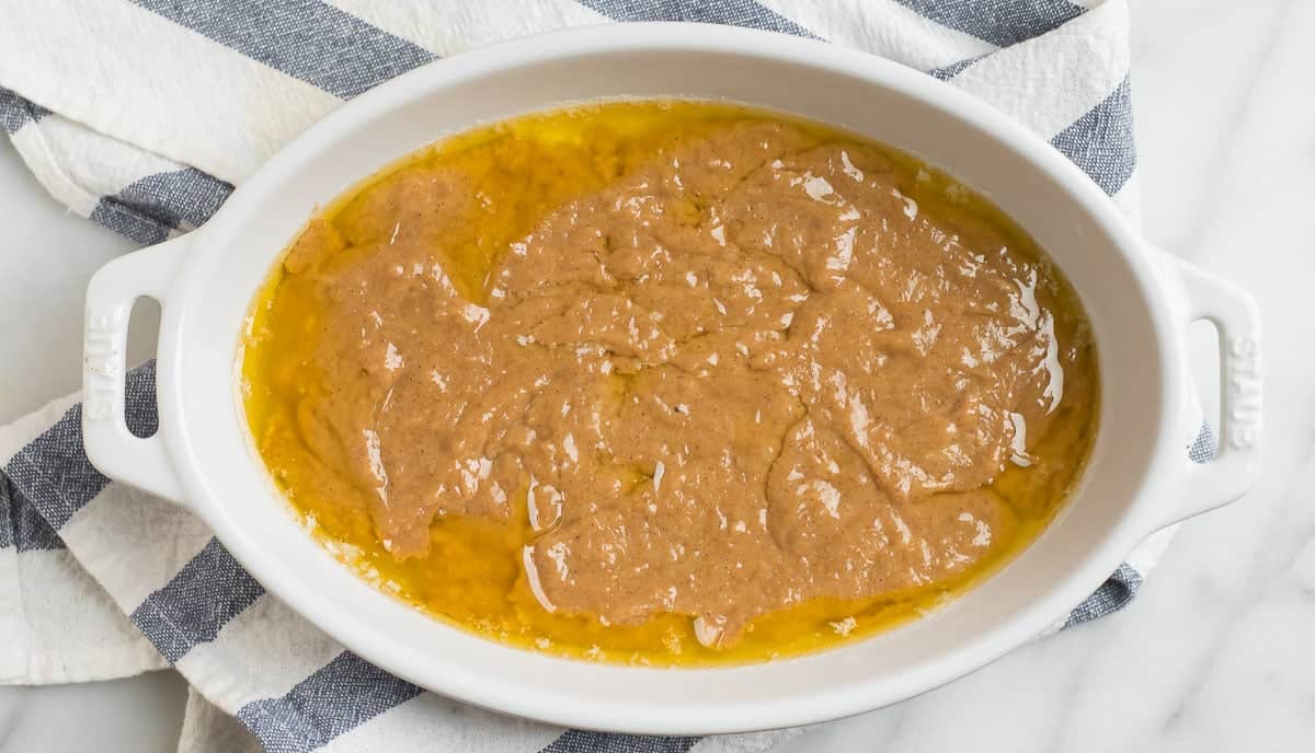 A white oval baking dish with a mixture of melted butter and brown batter on a striped kitchen towel.