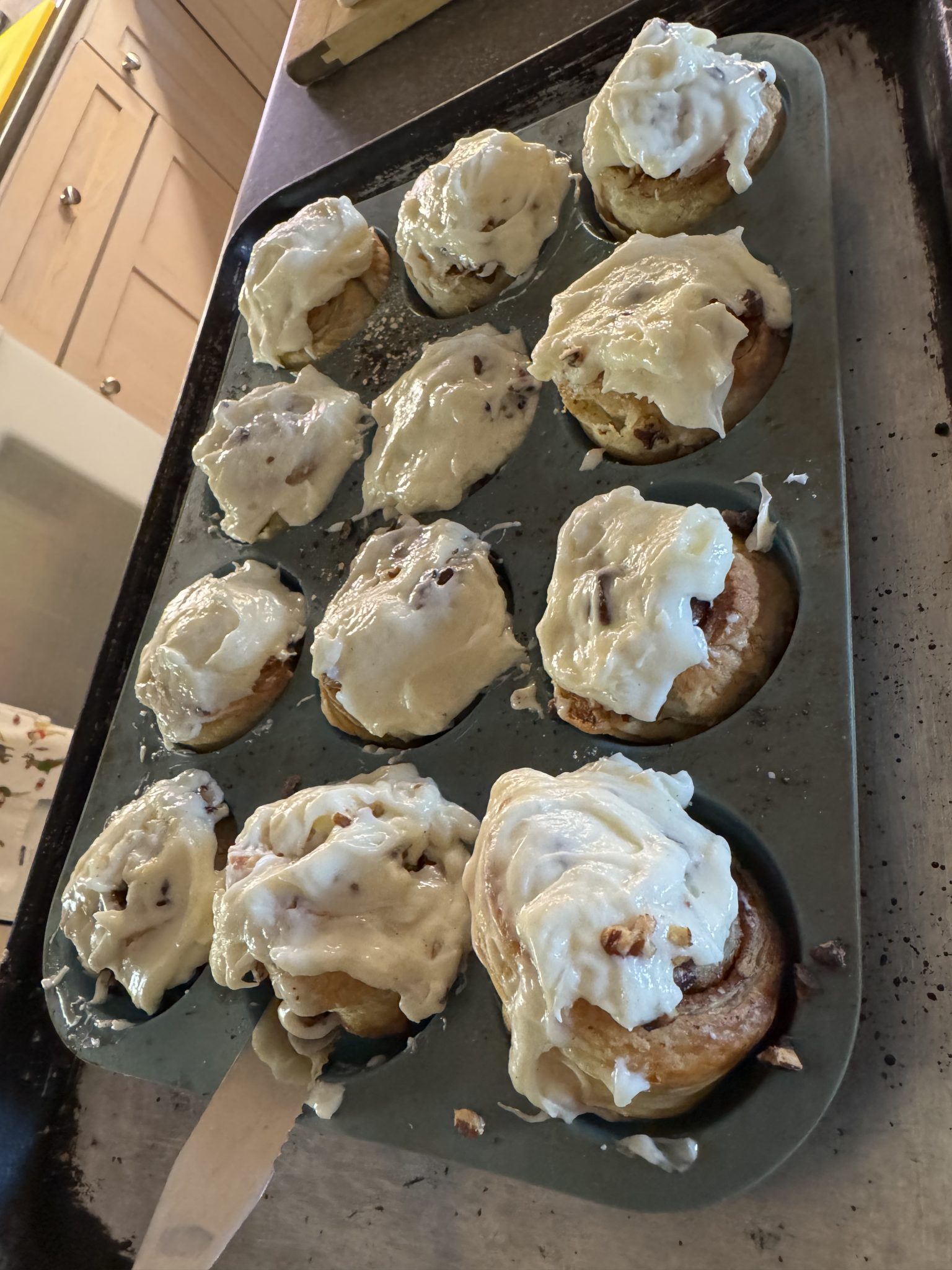 A tray of freshly baked cinnamon rolls with icing, some rolls overflowing from the muffin tin.