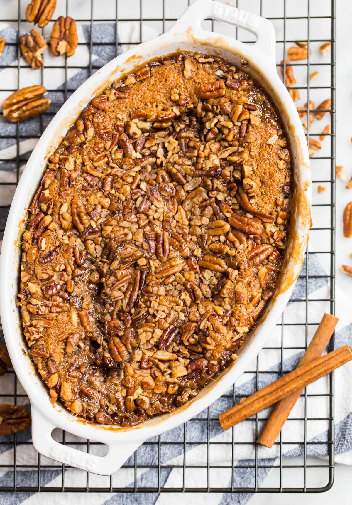 Oval baking dish filled with pecan-topped casserole, set on a cooling rack with cinnamon sticks and pecans nearby.