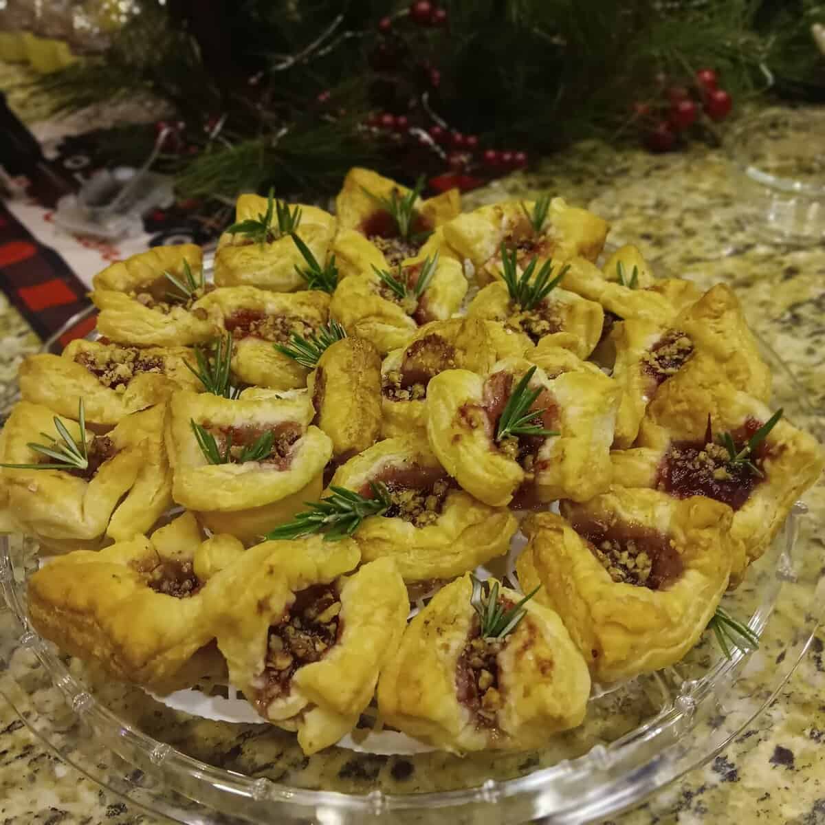 A platter of puff pastry bites filled with jam and nuts, garnished with fresh rosemary sprigs, on a festive table.