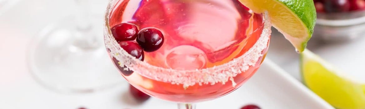 Two pink cocktails garnished with cranberries and lime, surrounded by cranberries and lime wedges on a white tray.