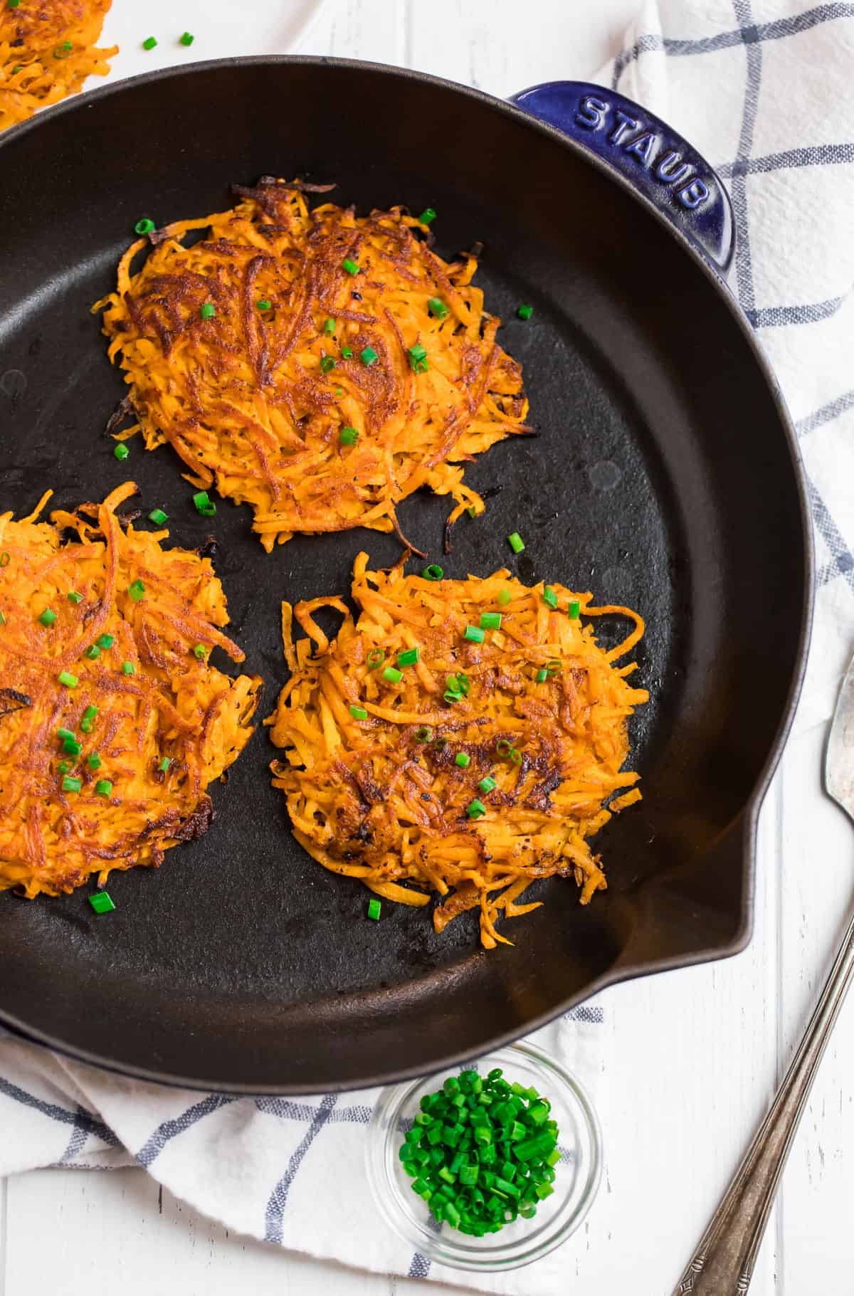 Three crispy sweet potato latkes topped with chopped chives in a black skillet, with extra chives in a small bowl nearby.