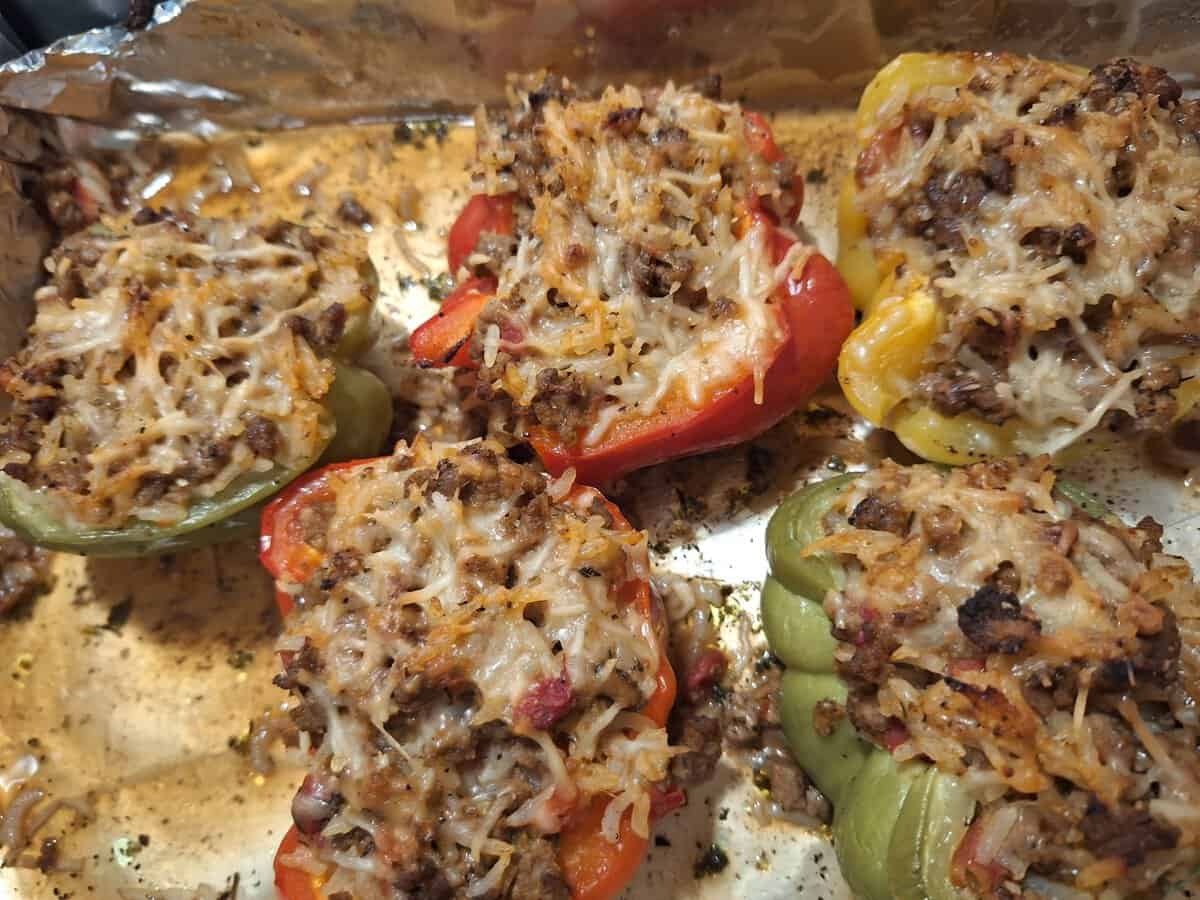 Five bell peppers, halved and stuffed with ground meat and rice, topped with melted cheese on a baking tray.