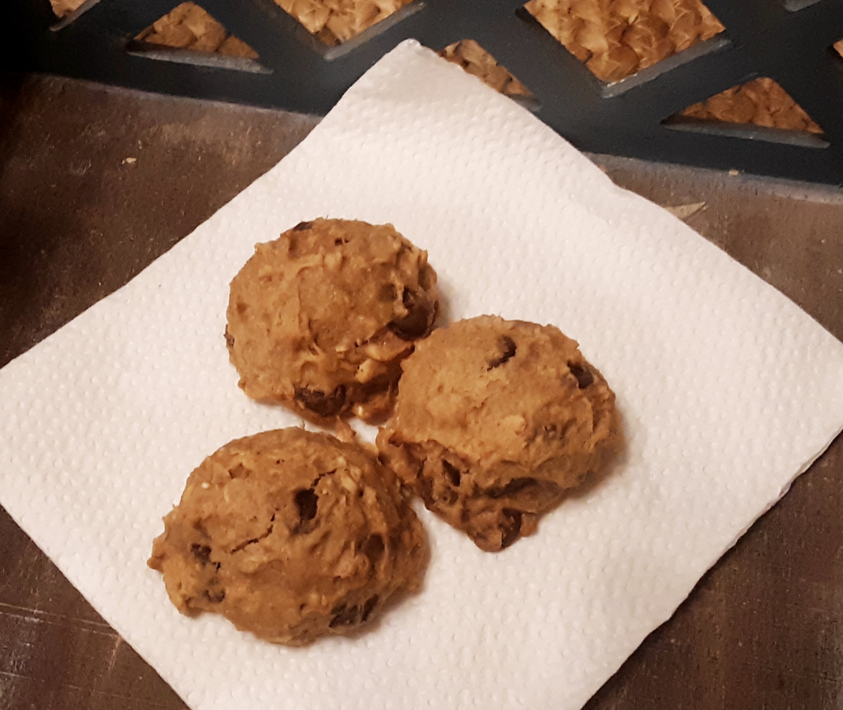 Three homemade cookies with chocolate chips on a white paper towel, placed on a wooden surface.