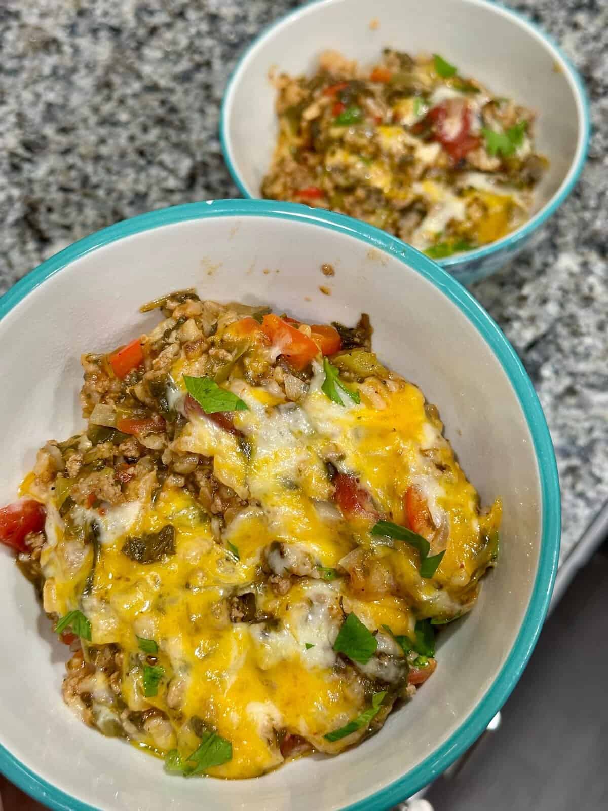 Two bowls of cheesy baked rice with vegetables, topped with melted cheese and fresh herbs, on a granite countertop.