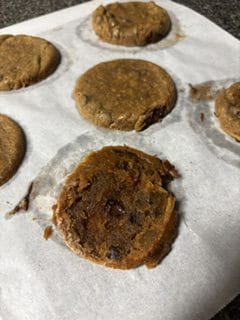 Six unevenly shaped cookies on parchment paper, with one cookie looking burnt and misshapen in the foreground.